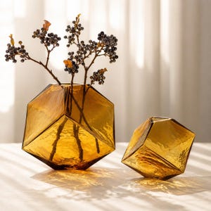 May include: Two amber-coloured glass vases with a geometric, multi-faceted shape. The larger vase contains several thin branches with small dark berries and a few dried leaves. The glass has a textured, translucent finish with sharp angles.