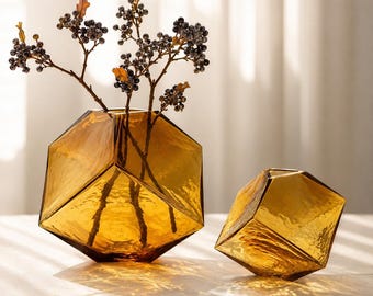 May include: Two amber-coloured glass vases with a geometric, multi-faceted shape. The larger vase contains several thin branches with small dark berries and a few dried leaves. The glass has a textured, translucent finish with sharp angles.