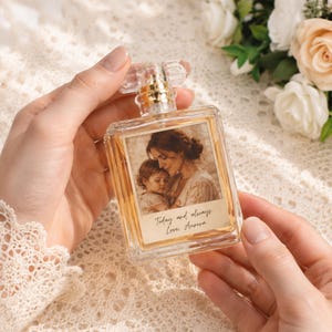 May include: A square glass perfume bottle filled with amber liquid, featuring a clear cap and a gold-toned spray nozzle. The front label displays a sepia-toned photograph of an adult and a child, with the text Today and always Love, Aurora.