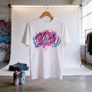 A white t-shirt with a custom graffiti-style design featuring the name "Olivier" in pink and blue colors, hanging on a wooden hanger in a studio setting.