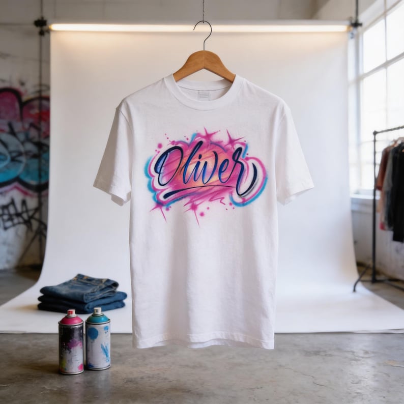 A white t-shirt with a custom graffiti-style design featuring the name "Olivier" in pink and blue colors, hanging on a wooden hanger in a studio setting.