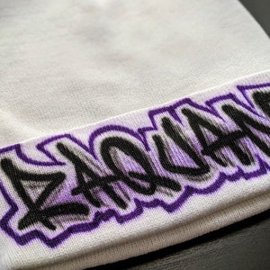Puede incluir: Gorro blanco con un diseño de graffiti morado y negro que dice "ZACHARY".