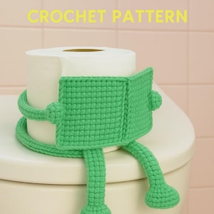 以下が含まれることがあります： 緑色のカギ針編みのトイレットペーパーカバー。カエル型で、上部に黄色の「CROCHET PATTERN」の文字。カエルは四角い頭、丸い目、長い足と球根状の足を持っています。カバーは白いトイレの上にあります。
