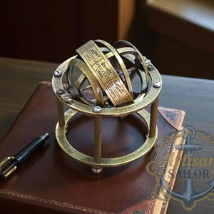 Puede incluir: Una esfera armilar de latón con signos del zodiaco, sobre un libro encuadernado en cuero. Un bolígrafo negro con detalles dorados está cerca. La esfera presenta anillos y una base, con una estética vintage.