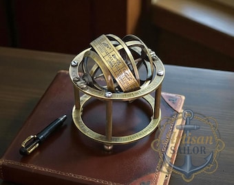 May include: A brass armillary sphere with zodiac signs, resting on a leather-bound book. A black pen with gold accents lies nearby. The sphere's design includes rings and a base, showcasing a vintage aesthetic.