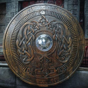 Puede incluir: Un gran escudo de madera redondo con intrincadas tallas. El escudo presenta un jefe central de metal y tallas en relieve de dragones entrelazados, nudos y runas. El escudo está teñido de un color marrón cálido, lo que sugiere un tema histórico o vikingo.