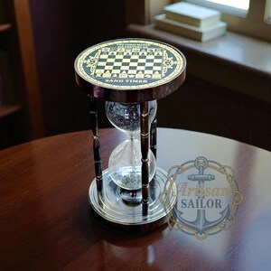 May include: A decorative hourglass sand timer with a chess board design on top. The timer has a dark wood frame with black accents and a clear glass chamber filled with sand. The base is silver-colored, and the text "SAND TIMER" is visible.