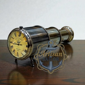 May include: A vintage-style clock in the shape of a telescope. The clock face is gold with black Roman numerals and the words "Victoria London 1901". The body is metallic with a brass finish. The clock is displayed on a dark brown surface.