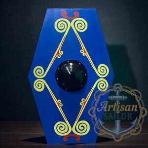 May include: A blue wooden shield with yellow and red decorative swirls and a central black boss. The shield has a hexagonal shape and is likely a decorative item or prop. The Artisan Sailor logo is visible in the lower right corner.