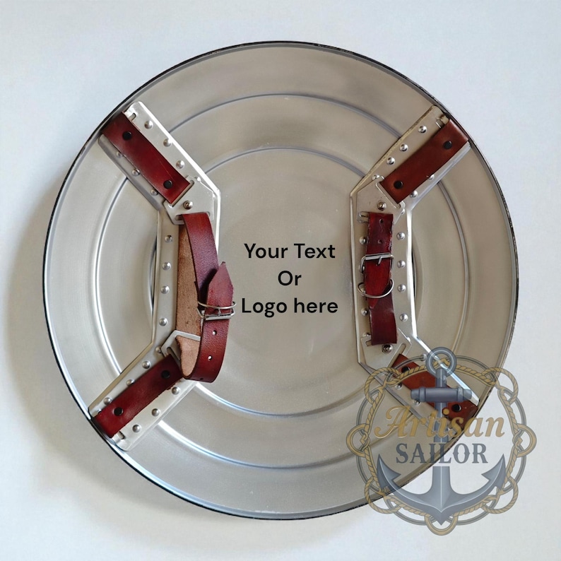 May include: A silver-colored, round shield with riveted metal supports and burgundy leather straps. The shield has the text "Your Text Or Logo here" in the center. The Artisan Sailor logo is in the lower right corner.