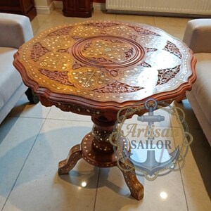 May include: A round, intricately carved wooden table with a dark brown finish. The table features detailed floral and geometric patterns, with a three-legged base. The table's diameter is approximately 30 inches.