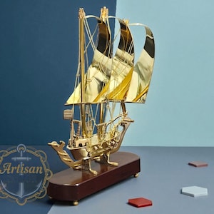 Könnte beinhalten: Ein goldfarbenes dekoratives Segelbootmodell mit großen, geschwungenen Segeln. Das Modell steht auf einem dunkelbraunen Holzsockel. Der Hintergrund ist zweifarbig blau. Kleine, farbige geometrische Formen sind um den Sockel verstreut.