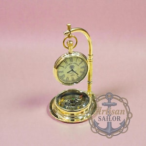 Può includere: Un set da scrivania in ottone con orologio e bussola. Il quadrante dell'orologio indica "Victoria London 1876" con numeri romani. La bussola è sotto l'orologio e il set è su una superficie rosa.