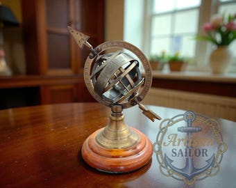 May include: A bronze-colored armillary sphere with an arrow design, mounted on a wooden and brass base. The decorative globe sits on a round, reddish-brown wooden base. The sphere is a vintage-style nautical decor item.