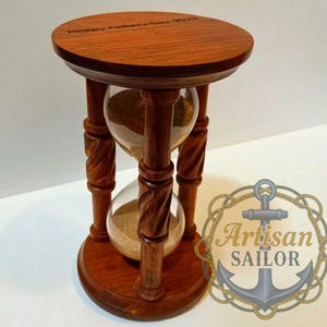 Può includere: Una clessidra in legno con finitura marrone scuro. La clessidra ha una parte superiore e inferiore rotonda, con un centro in vetro trasparente che contiene sabbia. La parte superiore della clessidra ha il testo "Happy Father's Day 2023".