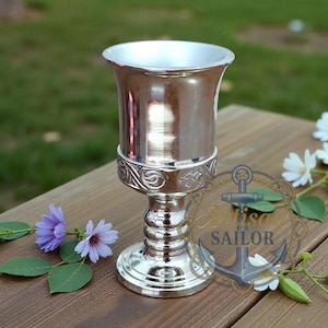 May include: A silver chalice with a decorative base and rim, placed on a wooden surface. The chalice has a flared top and an ornate design around the middle. Flowers and greenery are arranged around the chalice.