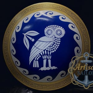 Puede incluir: Un escudo redondo con un centro azul que presenta un diseño de búho blanco y rama de olivo. El escudo tiene un borde dorado con un patrón repetitivo y un diseño de olas blancas. El logotipo de Artisan Sailor está en la esquina inferior derecha.