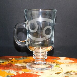 May include: Clear glass mug with a handle and a decorative base. The mug features the Cancer zodiac symbol etched on the front. The glass has a smooth surface and is designed for beverages.