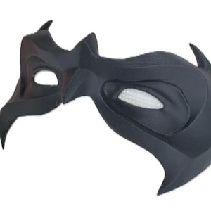 May include: A matte black mask with a sharp, angular design featuring pointed edges and a textured surface. The mask includes two eye openings, with one side displaying a white, grid-like mesh insert visible within the eye socket area.