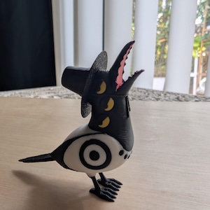 May include: A whimsical black, white, and yellow bird figurine with a top hat. The bird has a large eye design on its body and a beak with pink accents. The figurine is standing on a light-colored surface.