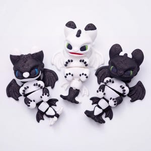 May include: Three dragon figurines in black and white. The dragons have large eyes, wings, and detailed features. One dragon is white with black accents, while the other two are black with white accents. The figurines are on a white background.