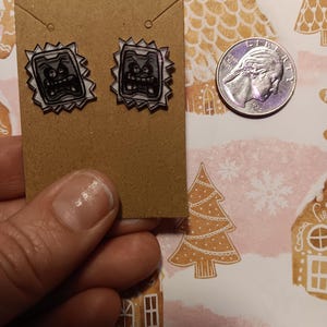 May include: A pair of black and white cartoon character earrings on a brown earring card. The earrings have a jagged border and a quarter is next to the earrings for size comparison. The background is a pink and white patterned paper with gingerbread house and tree designs.