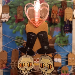 Puede incluir: Exhibición de pendientes hechos a mano, con diseños de calaveras y sombreros de copa. Los pendientes están sujetos a etiquetas marrones y suspendidos frente a un fondo pintado. Una lámpara rosa en forma de corazón está en la parte superior.