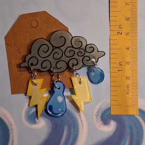 Op de afbeelding: Handgemaakte broche in de vorm van een wolk met bungelende bedels. De broche heeft een grijze wolk met zwarte contouren, gele bliksemschichten en blauwe regendruppelbedels. De liniaal meet 7.6 cm.