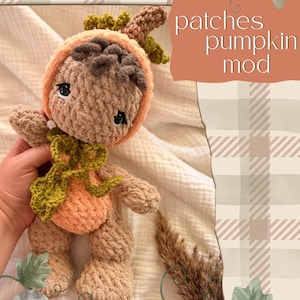 May include: A crochet doll with brown yarn hair, wearing a peach-colored pumpkin-shaped hood and a green crocheted collar. The doll has dark eyes and a soft, textured appearance. Text on the image reads: patches pumpkin mod.