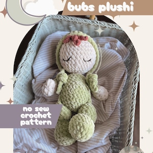 May include: A handmade green crochet plushie with a light green hood and a pink flower accent, resting in a woven basket. The text "bitty bubs plushie" and "no sew crochet pattern" are visible. The plushie is on a striped fabric.
