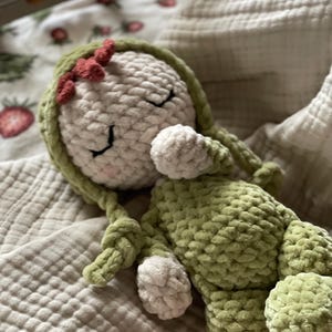 Puede incluir: Mu&ntilde;eca de ganchillo hecha a mano con cuerpo y cabeza verde claro y detalles blancos. La mu&ntilde;eca tiene los ojos cerrados, un peque&ntilde;o lazo rojo y largas trenzas verdes. Est&aacute; tumbada sobre una manta suave y texturizada.