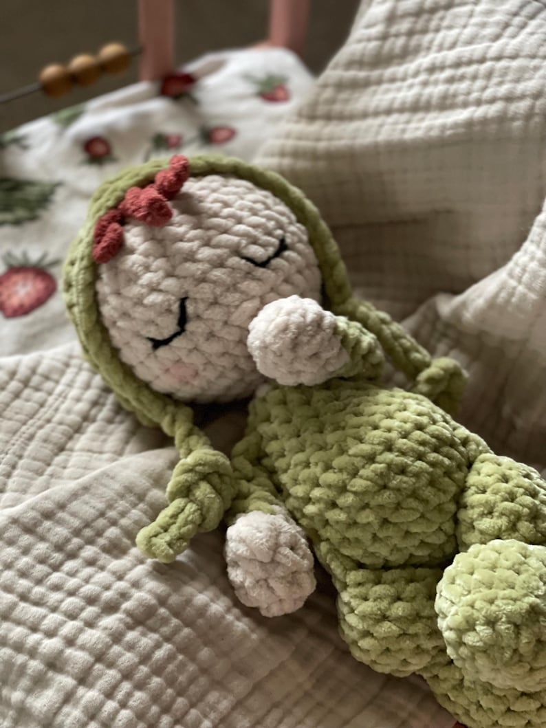 Puede incluir: Mu&ntilde;eca de ganchillo hecha a mano con cuerpo y cabeza verde claro y detalles blancos. La mu&ntilde;eca tiene los ojos cerrados, un peque&ntilde;o lazo rojo y largas trenzas verdes. Est&aacute; tumbada sobre una manta suave y texturizada.