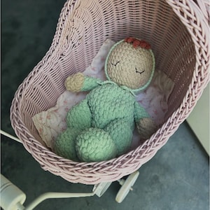 May include: A handmade crochet doll in a light green outfit and a beige bonnet with a red bow, resting in a pink wicker baby carriage. The carriage has white wheels and a white handle. The doll is lying on a floral patterned blanket.