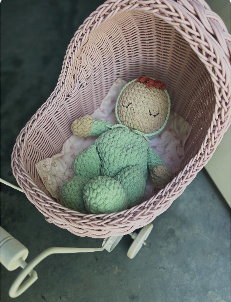 May include: A handmade crochet doll in a light green outfit and a beige bonnet with a red bow, resting in a pink wicker baby carriage. The carriage has white wheels and a white handle. The doll is lying on a floral patterned blanket.