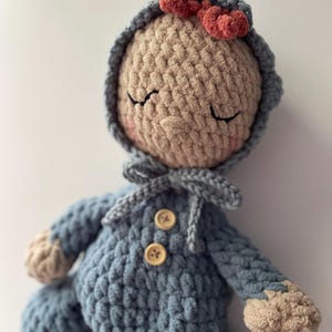 May include: A handmade crochet doll with a light blue outfit and a matching hood. The doll has a beige face with closed eyes, rosy cheeks, and a small red bow on the hood. The outfit features two wooden buttons.