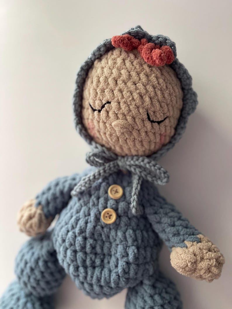 May include: A handmade crochet doll with a light blue outfit and a matching hood. The doll has a beige face with closed eyes, rosy cheeks, and a small red bow on the hood. The outfit features two wooden buttons.