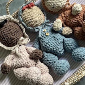May include: A collection of handmade crochet dolls in a woven basket. The dolls are in various colors, including blue, brown, and pink. Each doll has closed eyes and a unique outfit, with details like a bonnet and overalls.