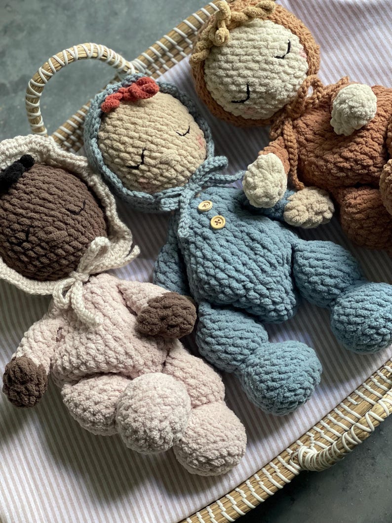 May include: A collection of handmade crochet dolls in a woven basket. The dolls are in various colors, including blue, brown, and pink. Each doll has closed eyes and a unique outfit, with details like a bonnet and overalls.