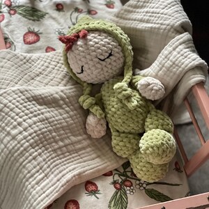Puede incluir: Una mu&ntilde;eca de ganchillo hecha a mano con un atuendo verde y una cabeza blanca, descansando en una cama de mu&ntilde;ecas rosa. La mu&ntilde;eca tiene un peque&ntilde;o lazo rojo y est&aacute; acostada sobre una manta con estampado de fresas. La cama est&aacute; forrada con una manta blanca y suave.