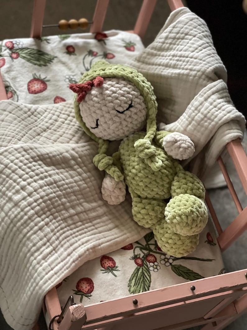 Puede incluir: Una mu&ntilde;eca de ganchillo hecha a mano con un atuendo verde y una cabeza blanca, descansando en una cama de mu&ntilde;ecas rosa. La mu&ntilde;eca tiene un peque&ntilde;o lazo rojo y est&aacute; acostada sobre una manta con estampado de fresas. La cama est&aacute; forrada con una manta blanca y suave.
