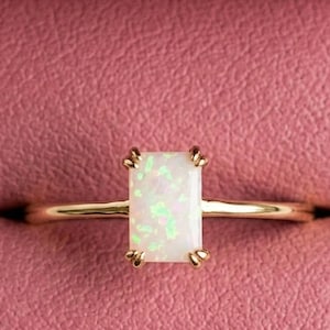 May include: A gold ring featuring a rectangular opal gemstone. The opal is milky white with green and yellow flashes. The ring is set in a simple style, and is presented in a pink jewellery box.