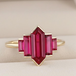 Könnte beinhalten: Ein goldener Ring mit einem zentralen, geometrisch geschliffenen, rubinroten Edelstein, flankiert von vier kleineren, rechteckigen Rubinen. Der Ring ist in einem goldenen Band gefasst und in einer rosa Schmuckschatulle präsentiert.
