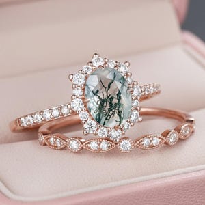 May include: A rose gold ring set featuring an oval-cut moss agate center stone surrounded by a halo of small round white stones. The band is accented with additional white stones, and a matching rose gold band with a milgrain edge is included.