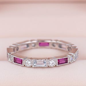 Puede incluir: Un anillo plateado con piedras preciosas rectangulares y redondas alternas. El anillo presenta piedras magenta y claras vibrantes, dispuestas en un patrón repetitivo. El anillo se muestra dentro de un joyero rosa.