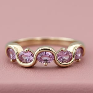 Puede incluir: Un anillo de oro con cinco piedras rosas ovales engastadas en un diseño ondulado. El anillo se muestra sobre un fondo rosa, resaltando el color de las piedras y el estilo elegante del anillo.