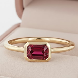 Peut inclure: Une bague en or avec une pierre précieuse rouge foncé rectangulaire sertie dans un sertissage à lunette. La bague est présentée dans un écrin rose. Le fond est de couleur neutre et douce.