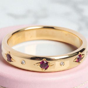 Pode incluir: Um anel de ouro com acabamento polido, apresentando três engastes em forma de estrela com pedras preciosas vermelhas escuras e intercaladas com pequenas pedras preciosas transparentes. O anel está sobre uma superfície rosa.