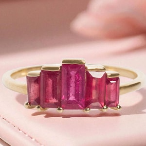 May include: A gold ring featuring five rectangular, vibrant pink gemstones. The ring's band is a polished gold, and the gemstones are set in a row across the top. The ring is displayed on a pink surface, creating a contrast with the gold and pink gems.