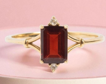 May include: A gold ring featuring a rectangular, deep red gemstone, flanked by two smaller diamonds. The ring's band has a delicate, openwork design. The setting is a warm gold color, and the background is a soft pink.