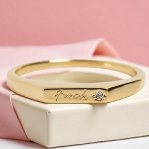 May include: A gold-colored ring with the word "fuck" engraved on the band. A small, clear gemstone is set next to the word. The ring is displayed on a white surface against a pink background.